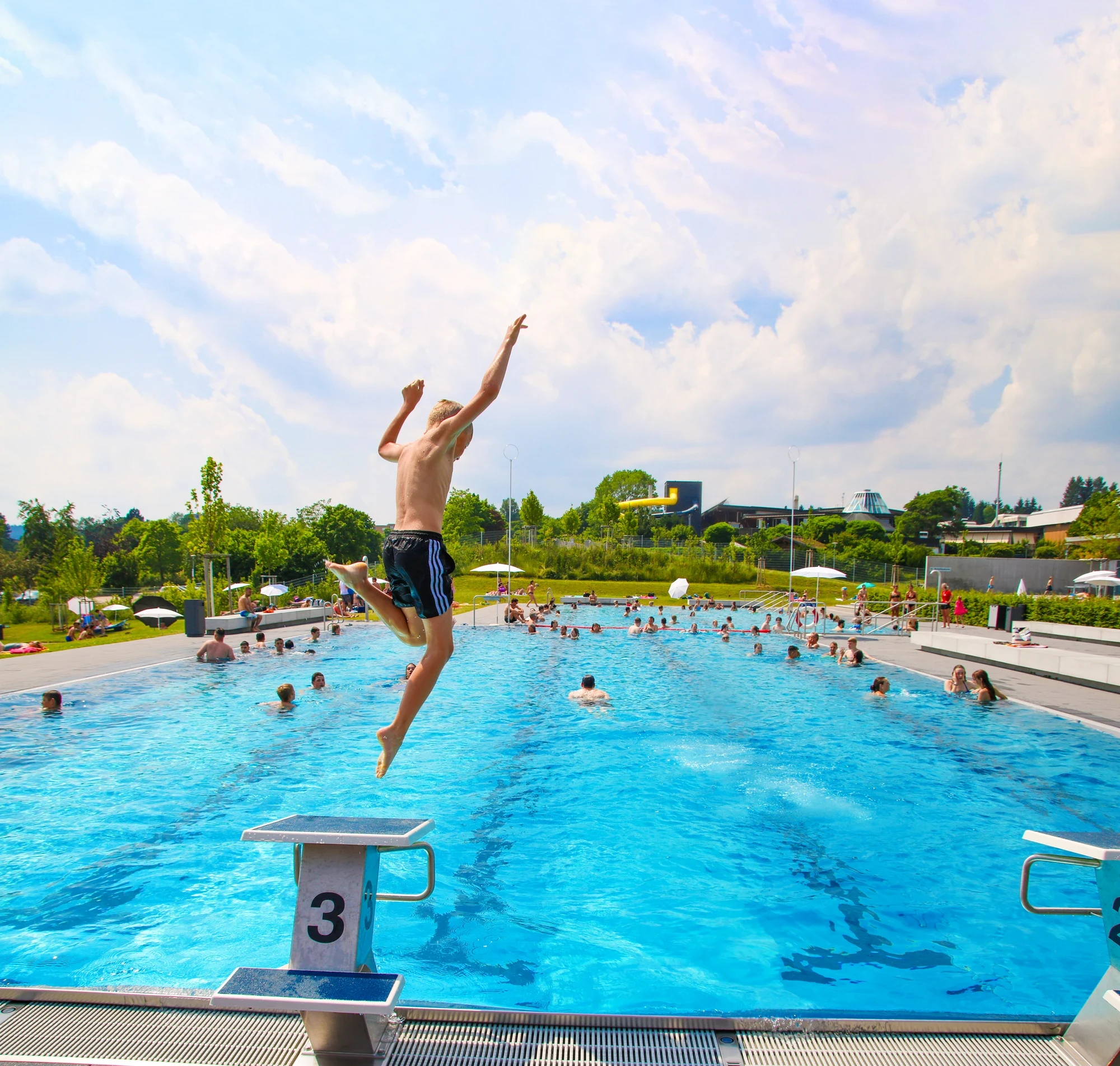 Junge springt vom Freibad-Startblock drei ins Wasser. Im Hintergrund zu sehen der Rutschenturm
