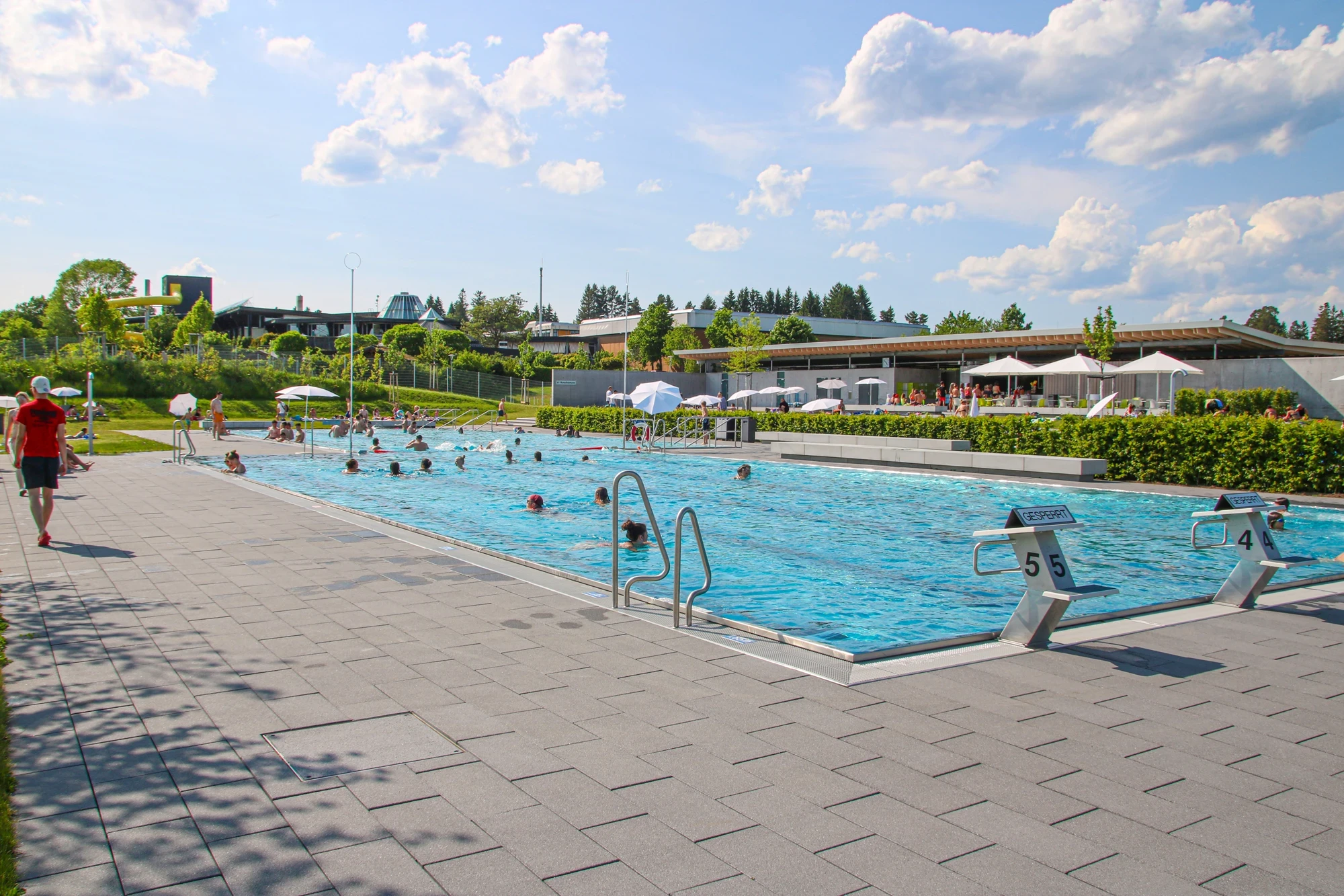 Freibad im Hintergrund der Kiosk mit seiner großen Terrasse mit Sonnenschirmen und dem Panorma-Bad mit Rutschenturm