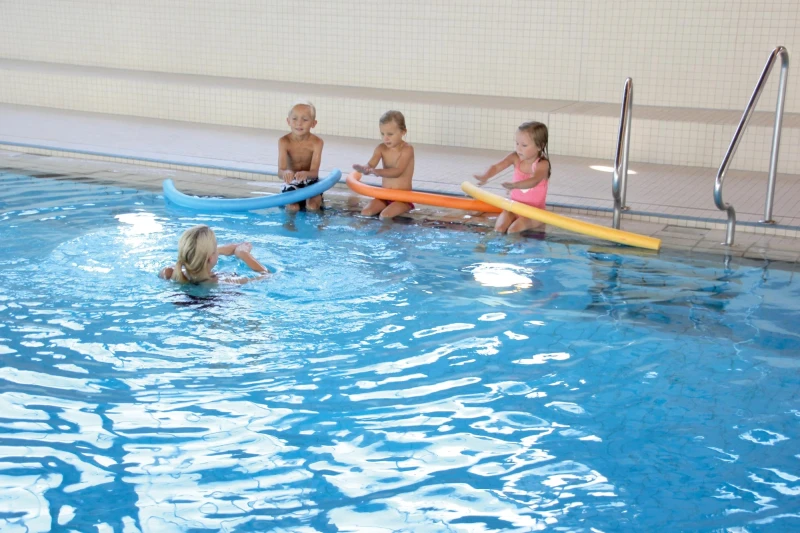 Schwimmtrainierin bringt Kindern am Beckenrand erste Schwimmtechniken mit bunten Poolnudeln bei