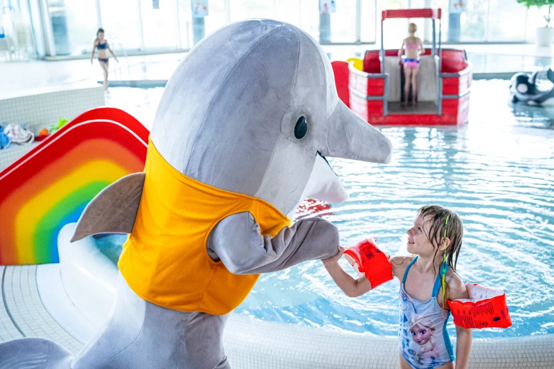 Maskottchen Flip, der Delfin, gibt einem Kind mit Schwimmflügeln beim Kinderplanschbecken die Hand